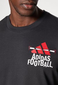 Sort bomulds t-shirt med et rødt og hvidt Adidas fodboldgrafik. Nærbillede af halsudskæringen og øverste brystområde, der fremhæver designet.