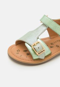 Sandalia metálica verde con un diseño de barra T, que incluye una hebilla dorada ajustable y una plantilla marrón texturizada adornada con patrones florales.