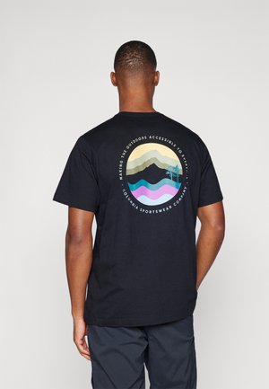 Uomo che indossa una maglietta nera con grafica colorata di montagna e albero e testo sull'accessibilità all'aperto sul retro, in piedi su uno sfondo semplice.