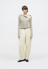 Bež dolg rokav s trakastim vzorcem in asimetričnim izrezom, kombiniran s kremnimi hlačami širokih hlačnic in črnimi ravnimi čevlji, na belem ozadju.