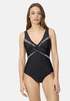 Mujer con un bañador entero negro con detalles cruzados estampados y tirantes anchos, de pie contra un fondo liso y claro.