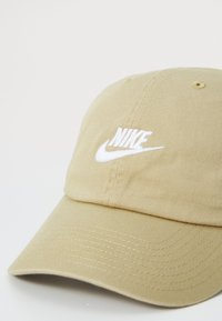 Beige baseball cap with a textured fabric, featuring a white embroidered Nike logo on the front and a curved visor.