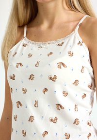 Biały top na ramiączkach z cienkimi paskami, ozdobiony wzorem małych brązowych wiewiórek i niebieskich kwiatów, noszony przez osobę o długich blond włosach.