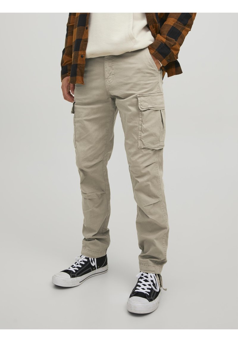 Personne portant un pantalon cargo beige, des baskets noires et blanches, un sweat-shirt blanc et une chemise à carreaux marron et noir avec les mains dans les poches.