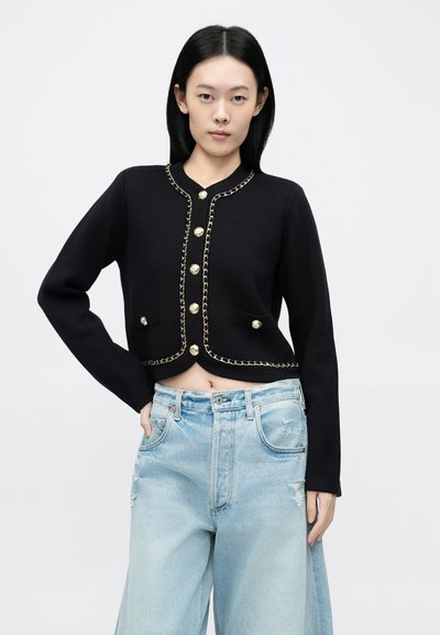 Fekete, kötött anyagból készült, rövidített kardigán arany gombokkal és lánc díszítéssel, világoskék, kopott, bő szárú farmerral párosítva.