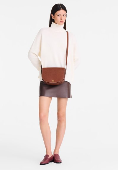 Braune Leder-Schultertasche mit einem gebogenen Klappdeckel, goldenen Hardware-Details, als Umhängetasche getragen. Modell in cremefarbenem Rollkragenpullover und brauem Lederrock, mit weinroten Schuhen.