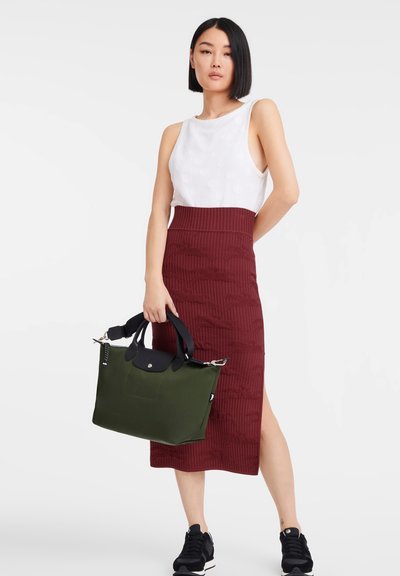 Green tote bag with black accents, textured burgundy pencil skirt, white sleeveless top, and black sneakers. Model stands with hand on hip.