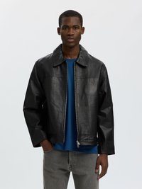 Junger Mann trägt eine schwarze Lederjacke über einem blauen Hemd und grauen Jeans, steht vor einem schlichten hellen Hintergrund.