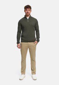 Ciemnozielony sweter z dzianiny z wysokim kołnierzem i suwakiem; teksturowany wzór. Dopasowany do beżowych spodni i białych sneakersów.
