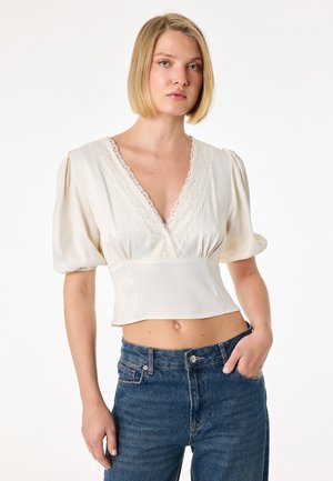 Kobieta o krótkich blond włosach nosi białą bluzkę z wycięciem w serek wykończoną koronką, z bufkami na rękawach oraz niebieskie dżinsy, pozując z jedną ręką w kieszeni.