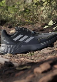 Čierno-šedá bežecká trailová obuv Adidas Terrex odpočívajúca na skalnatom vonkajšom povrchu so zeleným porastom v pozadí.