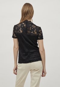 Svart spetsblus med korta ärmar, blommönster och en scalloped halsringning, kombinerad med krämfärgade byxor som framhäver en figurnära silhuett.