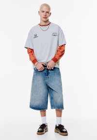 Grå grafisk t-shirt med grønne accenter lagdelt over en orange langærmet trøje. Afslappede blå denimshorts og sorte klodsede sko fuldender looket.