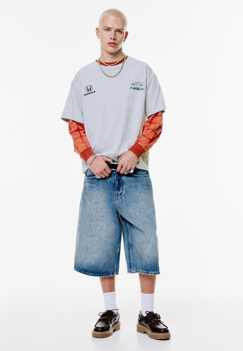 Grå grafisk t-shirt med grønne accenter lagdelt over en orange langærmet trøje. Afslappede blå denimshorts og sorte klodsede sko fuldender looket.