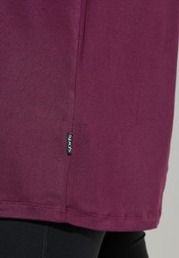 T-shirt de sport bordeaux avec une texture lisse, présentant une couture latérale et une petite étiquette noire "sports" près de l'ourlet. Conçu pour les vêtements de sport.