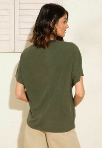Olivgrünes Strickoberteil mit kurzen Ärmeln, das einen lockeren Schnitt und ein dezentes, strukturiertes Muster aufweist. Der Rücken hat eine vertikale Nahtdetail.