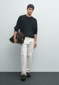 Mann in schwarzem Pullover, weißen Hosen und braunen Schuhen, der eine große braune Ledertasche gegen eine schlichte hellgraue Wand hält.