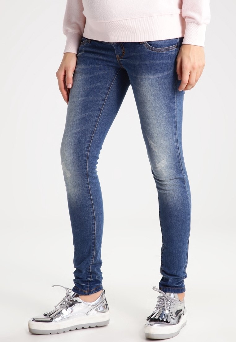 Femme portant un jean skinny bleu délavé et des chaussures à plateforme argentées brillantes à lacets, debout sur un fond blanc.