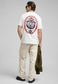 Vit bomullst-shirt med en stor röd och blå grafik på ryggen, med text och örndetaljer, kombinerad med beige byxor och svarta skor.