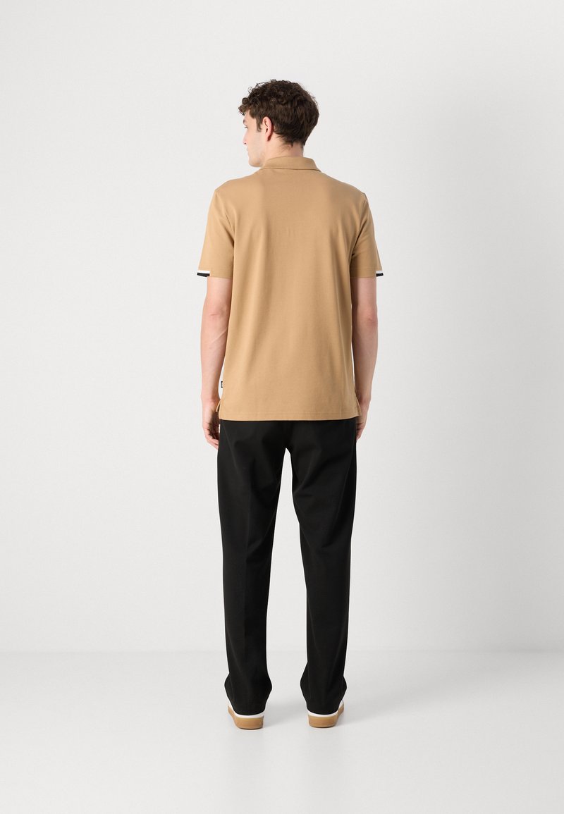 Polo shirt tan à manches courtes, avec des accents rayés noirs et blancs aux poignets, assorti à un pantalon noir et des semelles claires.
