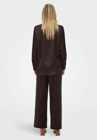 Femme aux longs cheveux blonds se tient de dos, portant un chemisier à manches longues marron foncé, un pantalon large assorti et des chaussures à talons carrés beige.