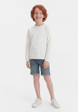 Garçon roux souriant debout, portant une chemise blanche à manches longues, un short en jean retroussé et des baskets blanches sur un fond blanc uni.