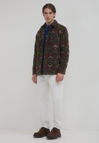 Un giovane uomo indossa una giacca a motivi verde scuro e bordeaux, una camicia blu, pantaloni bianchi e scarpe in suede marroni con lacci, mentre si trova contro uno sfondo semplice.