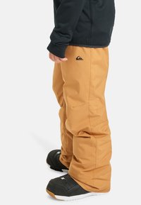 Svetlohnedé snehové nohavice so uvoľneným strihom, na prednej strane s čiernym logom. V páre s čiernymi snehovými topánkami a tmavým tričkom.