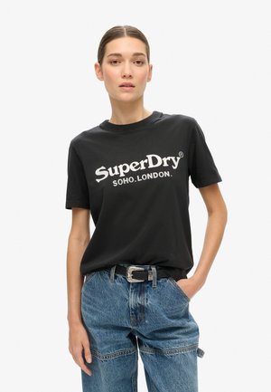 T-shirt en coton noir à manches courtes et col rond, arborant le logo blanc "SuperDry SOHO.LONDON." imprimé sur la poitrine. Jean en denim bleu.