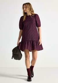 Kort lila klänning med texturerad tyg, puffärmar och volangkant. Kombinerad med vinröda strumpor och loafers, håller en vävd väska.