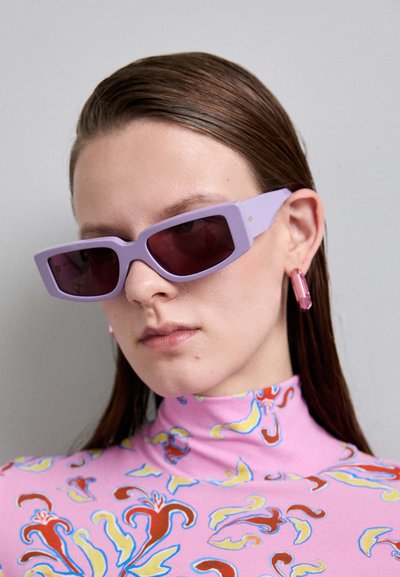 Lunettes de soleil rectangulaires violettes avec des verres teintés foncés, portées avec un col roulé rose à motifs présentant des designs floraux et des accents vibrants.