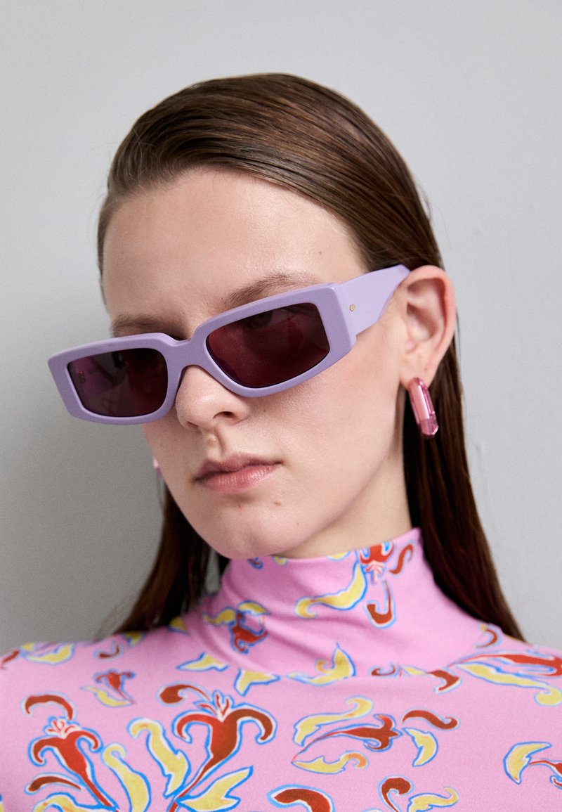 Occhiali da sole rettangolari viola con lenti scure, indossati con un dolcevita rosa a fantasia con motivi floreali e accenti vivaci.