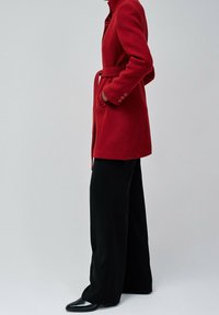 Abrigo de lana rojo con cuello, cinturón en la cintura y bolsillos laterales. Lleva puesto sobre pantalones negros de pierna ancha y zapatos negros puntiagudos. Diseño minimalista.