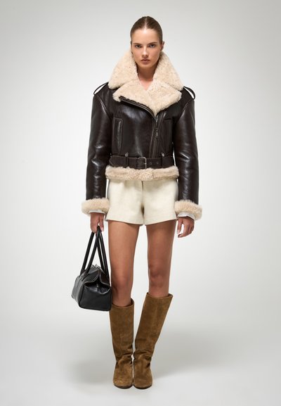 Jachetă de bomber din piele maro cu guler și manșete din blană crem. Asortată cu şorturi albe și cizme înalte din suede maro. Posetă neagră.