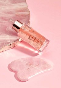 Serum do twarzy z różowego kwarcu w przezroczystej szklanej butelce z pipetą, wyposażonej w metalową srebrną zakrętkę. Towarzyszy mu narzędzie z różowego kwarcu w kształcie serca.