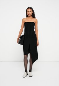 Melnā ribota bezplecu augšdaļa ar saišu detaļu, apvienota ar melnu asimetrisku svārku. Modelis valkā baltas slip-on kurpes un tur melnu rokassomu.