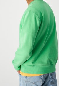 Person wearing a green sweater over a yellow shirt with hands in blue jeans pockets, turned slightly away on a plain white background.