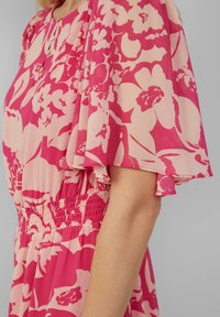 Robe à motifs floraux dans des tons de rose et rose clair, fabriquée en tissu léger. Elle présente des manches courtes et fluides ainsi qu'un détail à la taille froncée.
