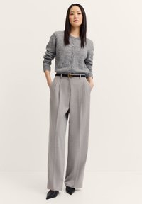 Grauer Wollcardigan mit Knöpfen, kombiniert mit hellgrauen, gefalteten, hochtaillierten Hosen und schwarzen spitz zulaufenden Absätzen. Glatte Textur und taillierte Passform.