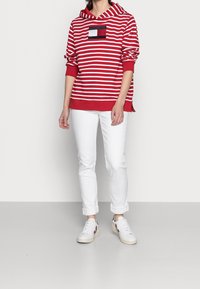 Hoodie a righe rosse e bianche con tasca frontale, abbinato a pantaloni bianchi e sneaker bianche. Il hoodie ha un fondo uniforme e maniche arrotolate.