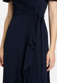 Robe portefeuille marine en tissu doux avec une ceinture nouée accentuant la taille. Caractérisée par des détails drapés et une texture lisse, mettant en valeur des lignes fluides.