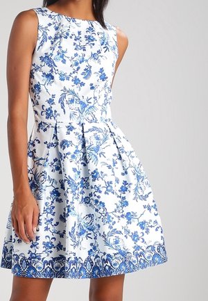 Mouwloze witte jurk met blauwe bloemen- en paisleypatronen, aansluitend lijfje en geplooide uitlopende rok, gedragen door een staande persoon.