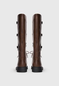 Stivali al ginocchio in pelle marrone con una finitura testurizzata, caratterizzati da sei bottoni decorativi e lacci annodati sul retro, su una suola in gomma nera.