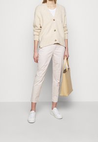 Un cardigan en laine beige avec trois boutons, associé à un pantalon beige clair. Des baskets blanches et un sac fourre-tout jaune complètent la tenue.