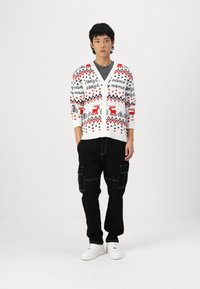 Hvid cardigan med røde rensdyr og sorte mønstre, knaplukning, båret over en mørk T-shirt. Sorte cargo-bukser og hvide sneakers fuldender outfittet.