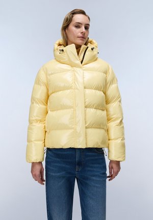 Person iført en skinnende lysegul dunjakke med høj krave og hætte, kombineret med blå jeans, stående mod en ensfarvet baggrund.