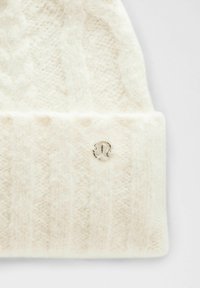 Valkoinen neulepipo, joka on valmistettu pehmeästä materiaalista ja jossa on kaapelikuvio. Pipossa on pieni hopeinen logo yksityiskohta käännetyssä reunassa.