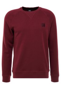 Sudadera burdeos con un cuello redondo clásico, puños y dobladillo acanalados, y una pequeña etiqueta de logo negra en el pecho izquierdo. Textura suave.