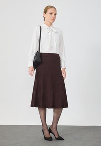 Blouse blanche avec un nœud et des manches longues, associée à une jupe midi plissée noire et des escarpins noirs. Un petit sac à main noir est porté en bandoulière.