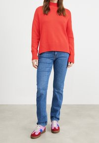 Červený pletený svetr s vysokým límcem, zkombinovaný s modrými džíny rovného střihu a multikolorovými teniskami s růžovými, červenými a modrými akcenty.